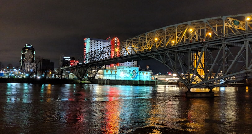 A brightly lit casino with neon signs and red lights is reflected in a river. A bridge stretches across the water, with an urban skyline visible in the background against an overcast night sky.
